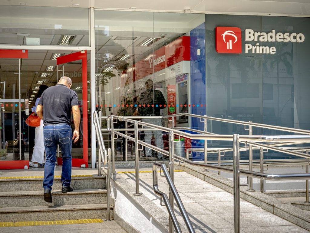Homem sobe escadas na entrada de agência Bradesco Prime, que possui rampa de acessibilidade e corrimãos.