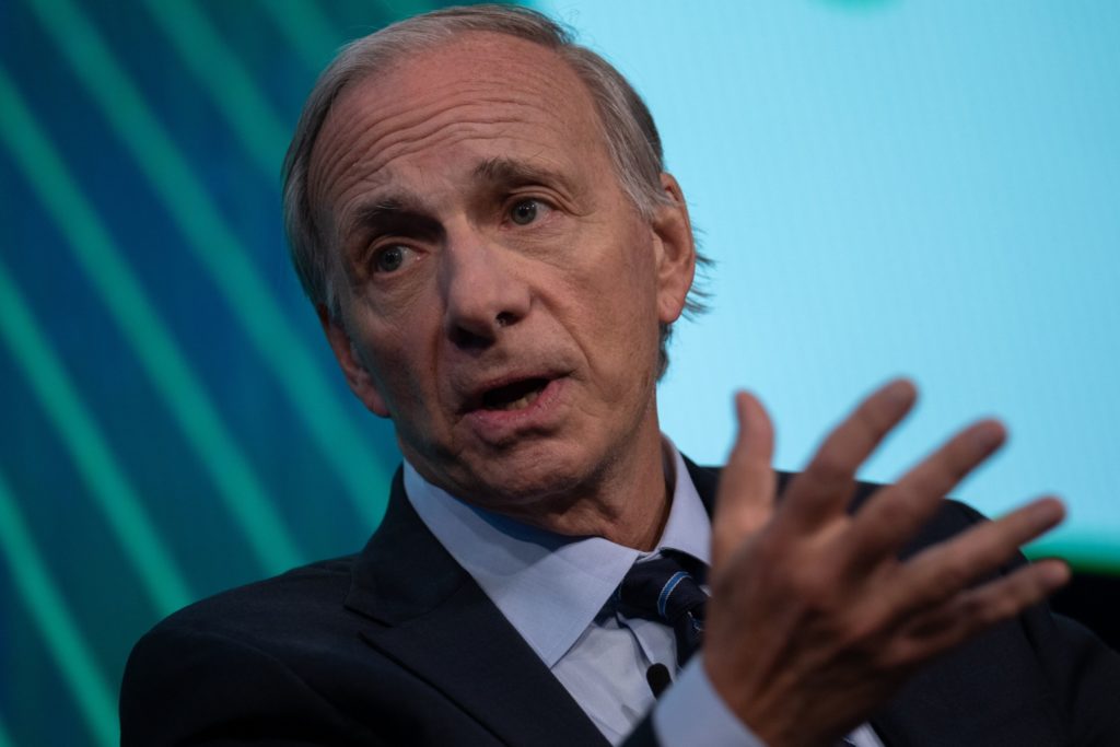 Ray Dalio, de terno e gravata, fala gesticulando com a mão aberta. Fundo azul.