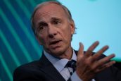 Ray Dalio, de terno e gravata, fala gesticulando com a mão aberta. Fundo azul.