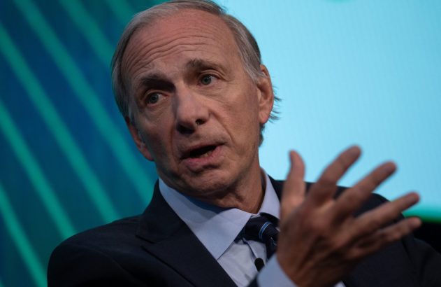 Ray Dalio, de terno e gravata, fala gesticulando com a mão aberta. Fundo azul.