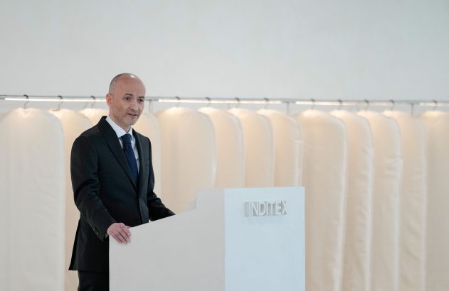 Homem de terno em pódio INDITEX, falando. Capas de roupa brancas penduradas ao fundo.