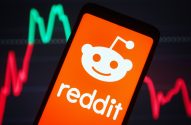 Smartphone com logo do Reddit. Gráficos de mercado financeiro (verde e vermelho) desfocados no fundo.