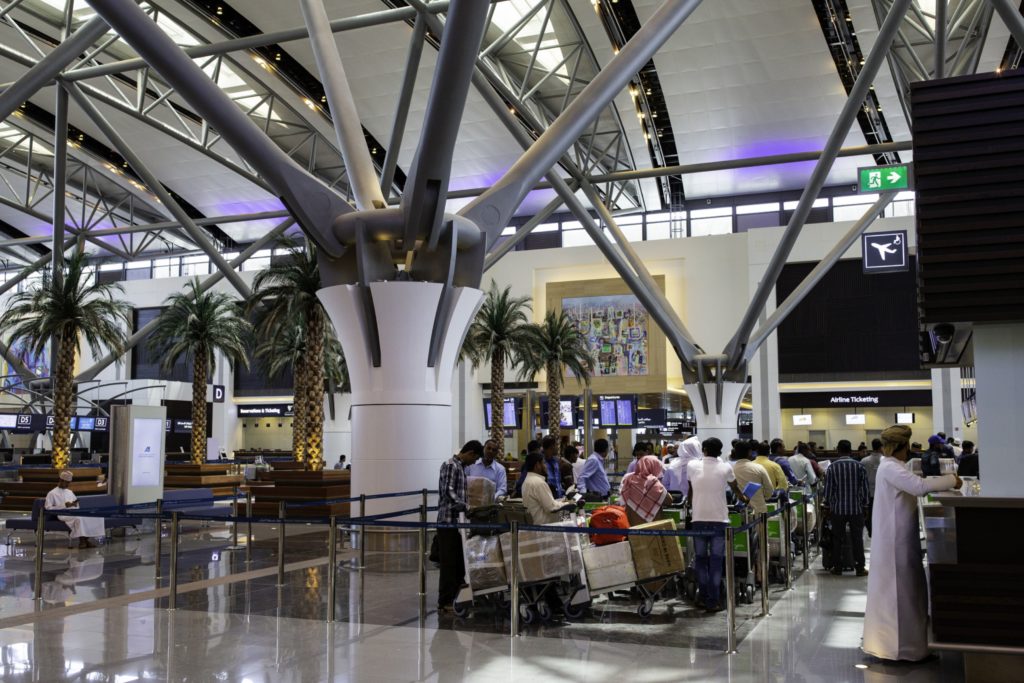 Pessoas em filas com bagagem em um terminal de aeroporto moderno com grandes colunas estruturais e palmeiras.