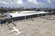 Aeroporto com terminal moderno e aviões no pátio. Ao fundo, skyline urbano sob céu nublado.