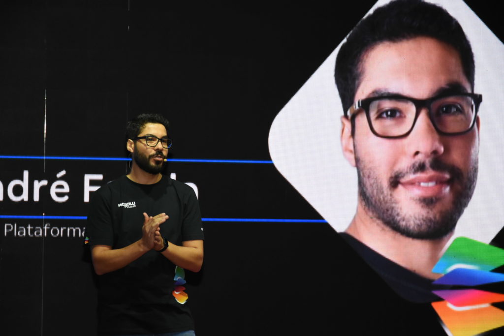 Homem de óculos em palco apresenta, com tela exibindo seu rosto e nome 'André F...'.
