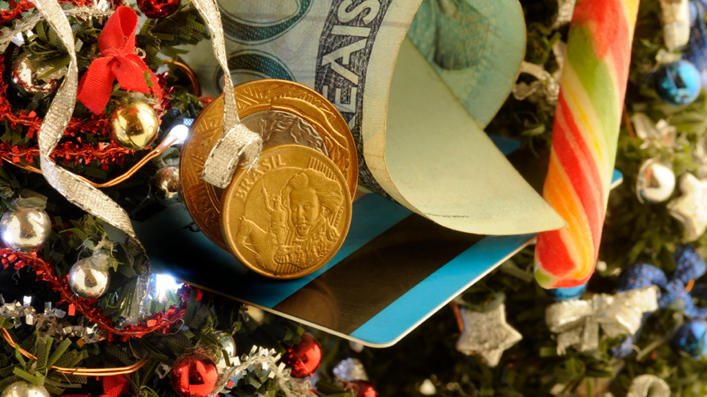 Notas, moedas, cartão e um doce natalino sobre uma árvore de Natal decorada com bolas e laços.