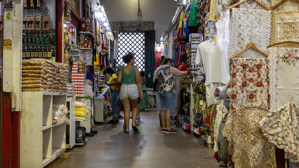 Corredor de mercado interno. Pessoas compram em bancas com alimentos, bebidas, roupas, tecidos e artesanato.