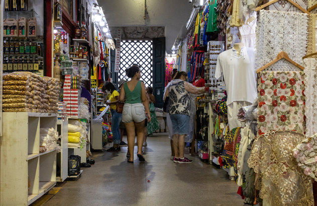 Corredor de mercado interno. Pessoas compram em bancas com alimentos, bebidas, roupas, tecidos e artesanato.