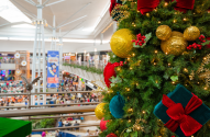 Árvore de Natal decorada em primeiro plano com shopping center movimentado e pessoas ao fundo.