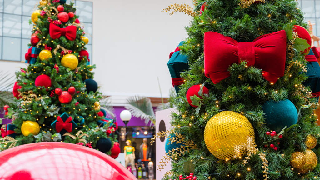 Duas árvores de Natal verdes decoradas com laços vermelhos, bolas douradas e luzes brilhantes, em ambiente interno.
