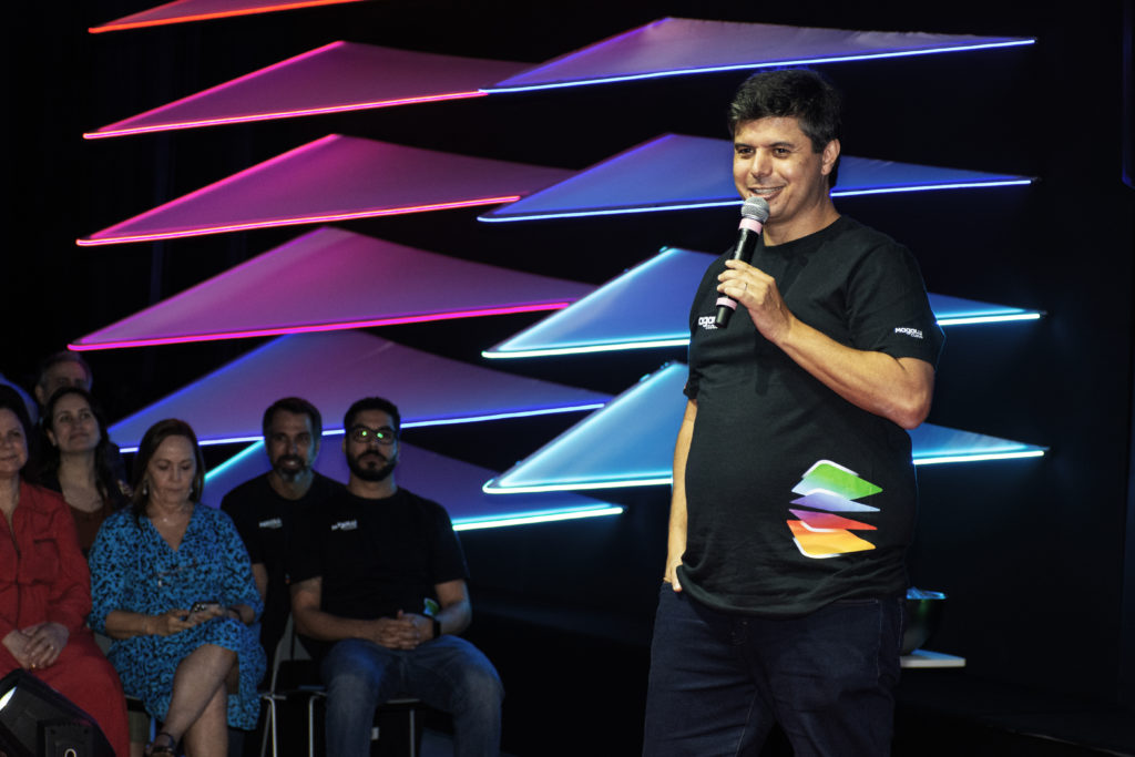Homem sorrindo com microfone no palco, apresentando. Fundo com triângulos luminosos coloridos e audiência.