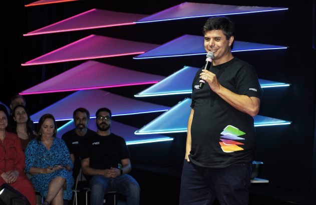 Homem sorrindo, falando ao microfone em palco. Fundo com design moderno de luzes triangulares em tons neon.