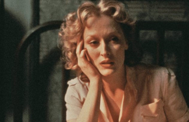 Meryl Streep com cabelo loiro cacheado, expressão pensativa, apoiando o rosto na mão, em cena escura com luz quente.
