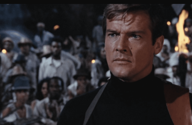 Homem de meia gola preta com alça no ombro, olhando sério, entre multidão e tochas à noite.