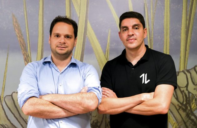 Dois homens em pé, braços cruzados, olhando a câmera. Um em camisa azul claro, outro em polo preta.