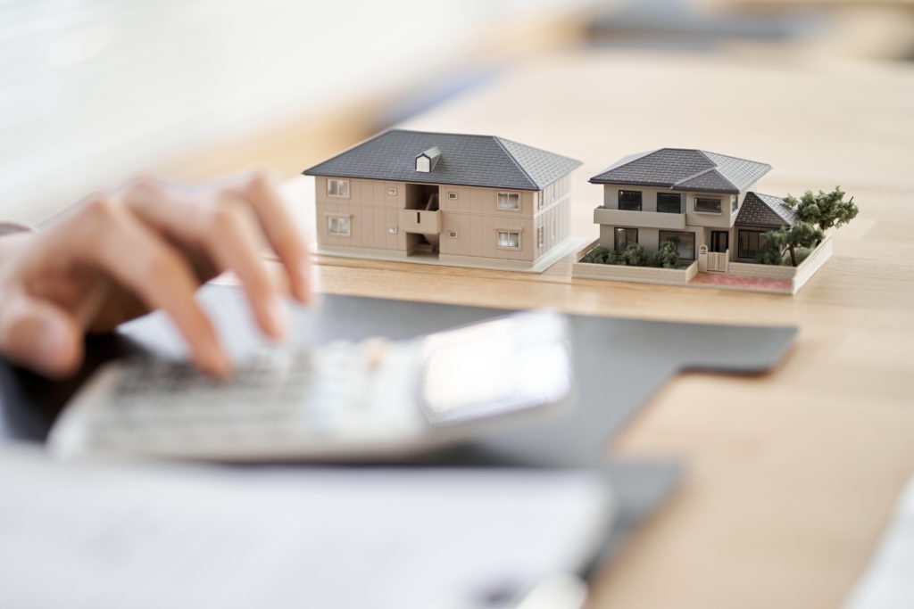 Mão digitando em calculadora, com dois modelos de casas em miniatura sobre mesa de madeira.