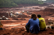 Duas pessoas observam uma vasta área de terra vermelha com maquinário, sugerindo mineração ou desastre ambiental.