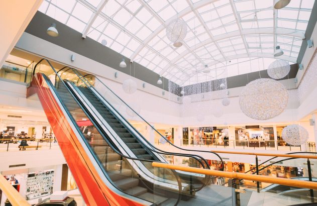 Interior de shopping iluminado, com escadas rolantes, lojas em vários andares e um grande teto de vidro. Luzes decorativas.
