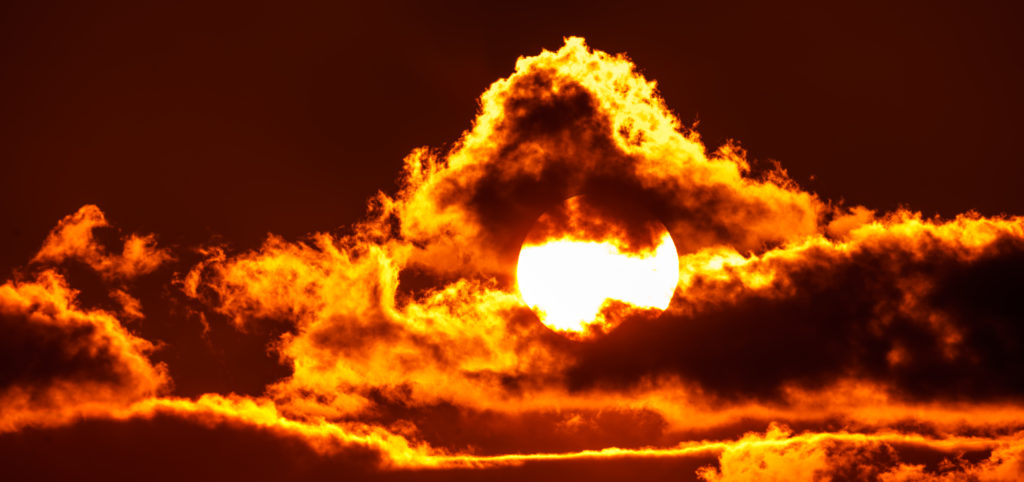 O sol irradia luz intensa através de nuvens alaranjadas em um céu escuro.