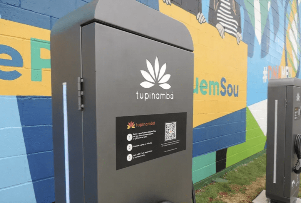 Carregador de veículo elétrico Tupinambá com QR code e instruções, ao lado de um mural colorido.