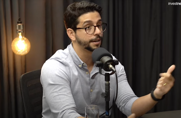 Homem de óculos e camisa clara falando em microfone, gesticulando em um podcast.