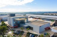 Vista aérea de complexo industrial moderno, com escritórios e galpões. Dia ensolarado.