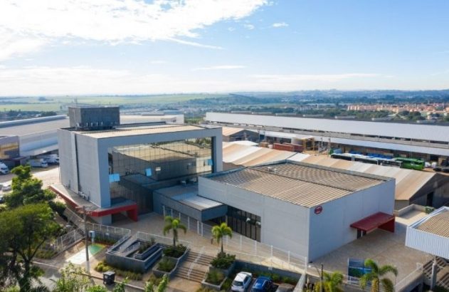 Vista aérea de complexo industrial moderno, com escritórios e galpões. Dia ensolarado.