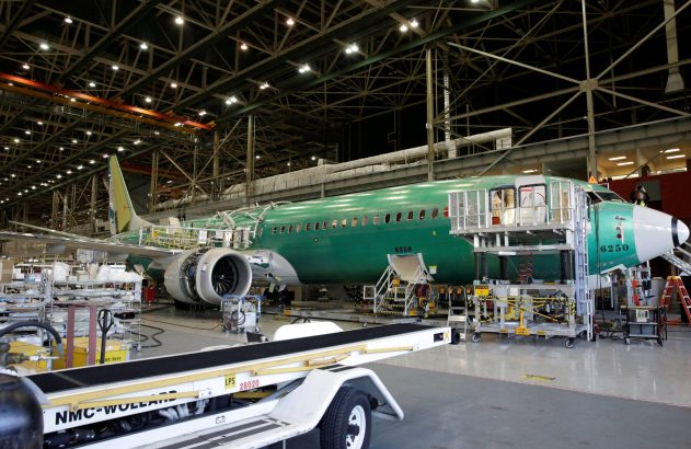 Fuselagem verde de avião em construção dentro de um hangar, cercada por equipamentos.