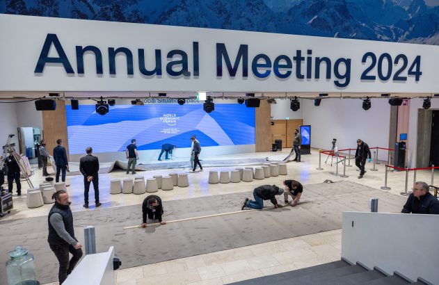 Pessoas preparam salão para o Fórum Econômico Mundial 2024, instalando piso sob letreiro 'Annual Meeting 2024'.