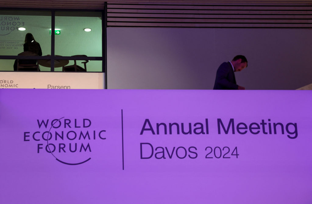 Banner do Fórum Econômico Mundial Davos 2024 com homem de terno em pé acima e outras pessoas ao fundo.