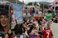 Fãs sentados em grupo em área externa, aguardando evento. Há um banner de Taylor Swift à esquerda.