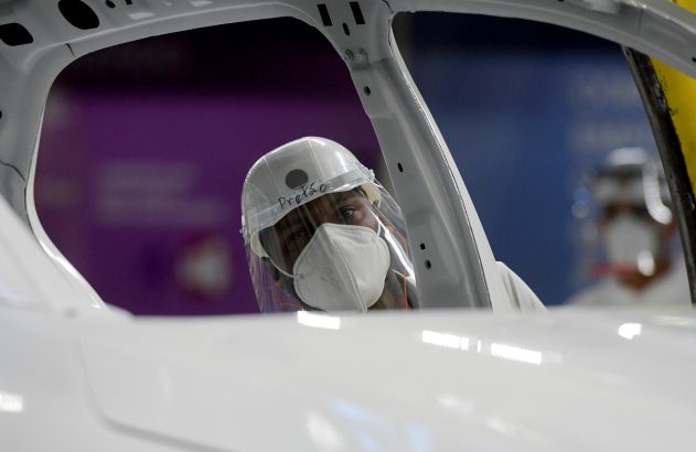 Pessoa com máscara e capacete trabalhando dentro da carroceria branca de um carro em fábrica.