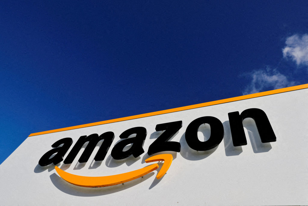 Vista de baixo de uma placa da Amazon com o logotipo da seta laranja e céu azul com nuvens.