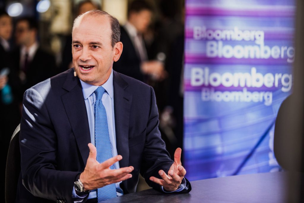 Homem de terno e gravata, com microfone, fala e gesticula em estúdio com logos Bloomberg.