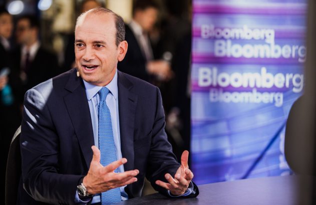 Homem de terno e gravata, com microfone, fala e gesticula em estúdio com logos Bloomberg.