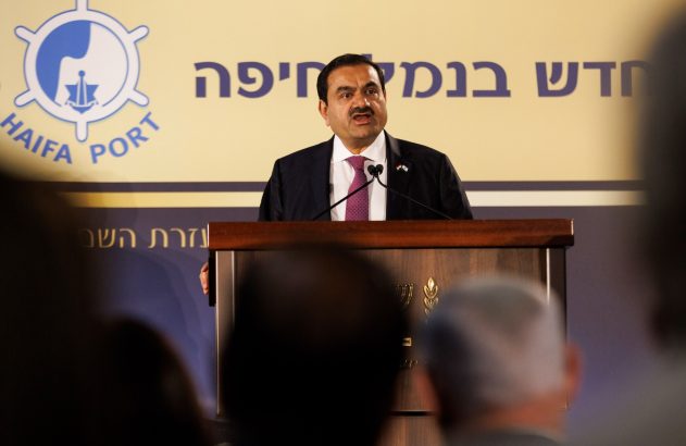 Homem de terno discursa em pódio, com logo do Porto de Haifa e texto em hebraico ao fundo.