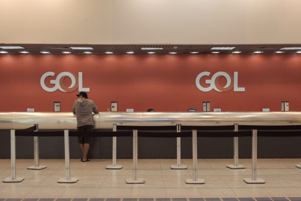 Pessoa em balcão de atendimento da Gol, com barreiras de fila. Fundo vermelho com logo Gol.