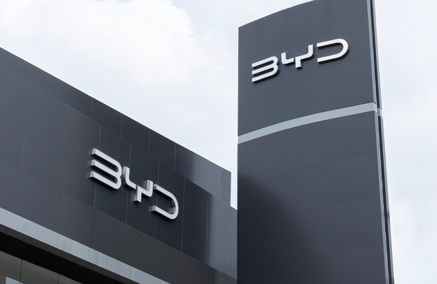 Prédio e totem modernos da BYD. Dois logos da marca em relevo na fachada cinza escura e no totem contra o céu.