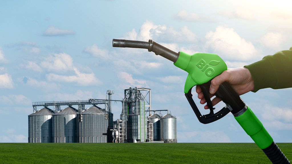 Mão segura bico de abastecimento verde com 'BIO'. Silos de grãos em campo verde sob céu azul.