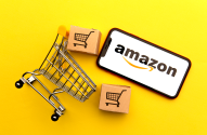Conceito de e-commerce: carrinho de compras, caixas e smartphone com logo Amazon em fundo amarelo.