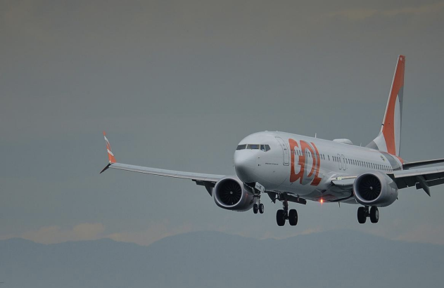 Avião GOL branco e laranja descendo com trem de pouso, torre de igreja à esquerda, sob céu nublado.