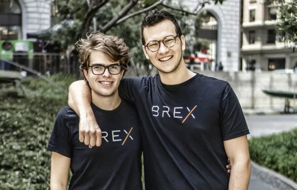 Dois homens sorrindo, ambos de óculos e camisetas pretas com o logo BREX, um com braço no ombro do outro, ao ar livre.