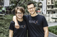 Dois homens sorrindo, ambos de óculos e camisetas pretas com o logo BREX, um com braço no ombro do outro, ao ar livre.