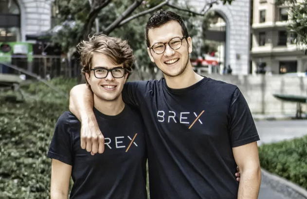 Dois homens sorrindo, ambos de óculos e camisetas pretas com o logo BREX, um com braço no ombro do outro, ao ar livre.