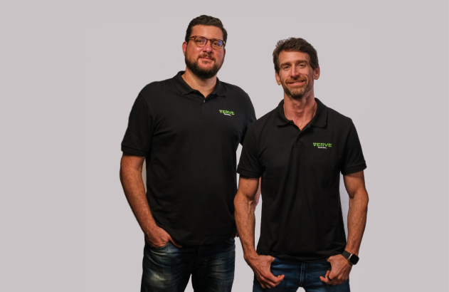 Dois homens de pé com camisas polo pretas e jeans, olhando para a câmera em fundo neutro.