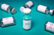Frascos de vacina COVID-19 sobre superfície turquesa, com um em destaque no centro.