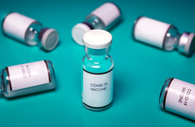 Frascos de vacina COVID-19 sobre superfície turquesa, com um em destaque no centro.