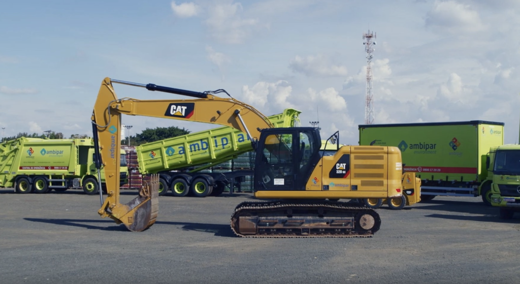 Escavadeira CAT 320 GC amarela em pátio, braço elevado sobre caminhão Ambipar, outros veículos verdes atrás.