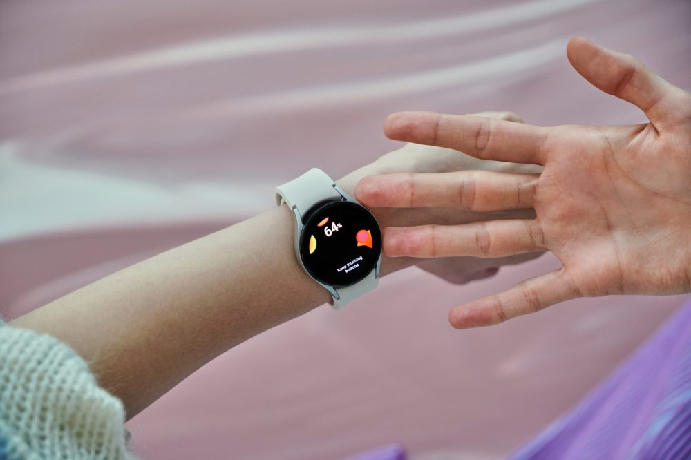 Mão usa smartwatch branco com tela de ícones coloridos, outra mão aberta ao lado.