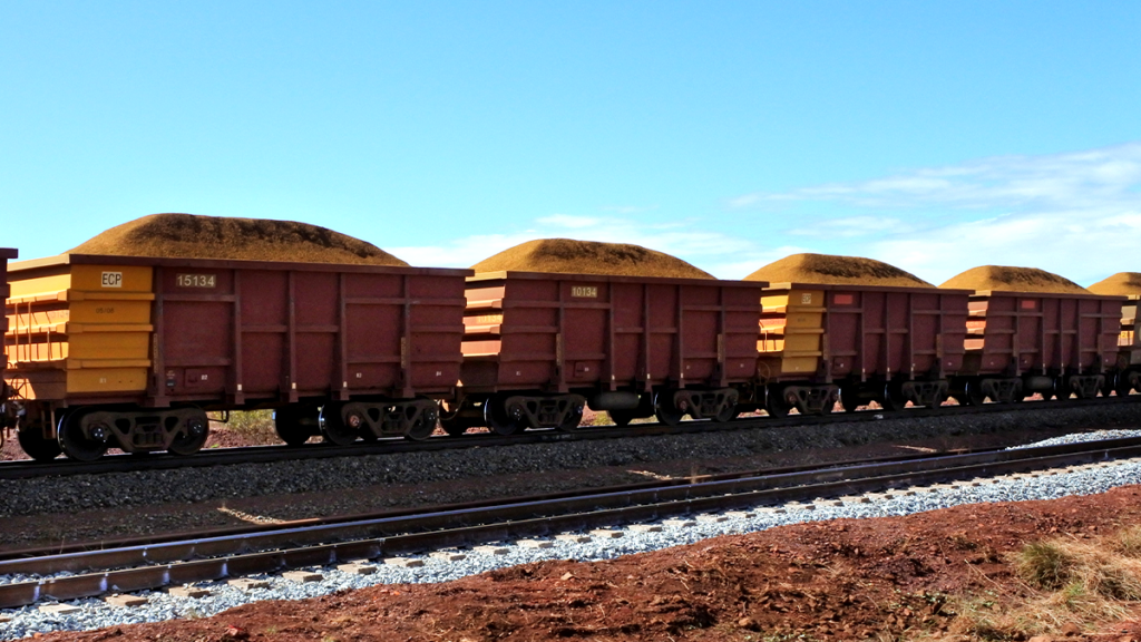 Vagões de trem carregados de minério em trilhos sob céu azul. Terreno avermelhado.